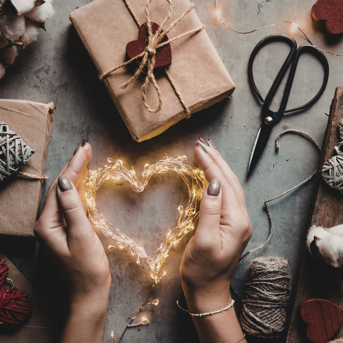A top-down flat lay of hands holding a glowing heart-shaped string light, surrounded by wrapped brown paper gift boxes, scissors, twine, cotton bolls, and small decorative hearts on a gray surface.